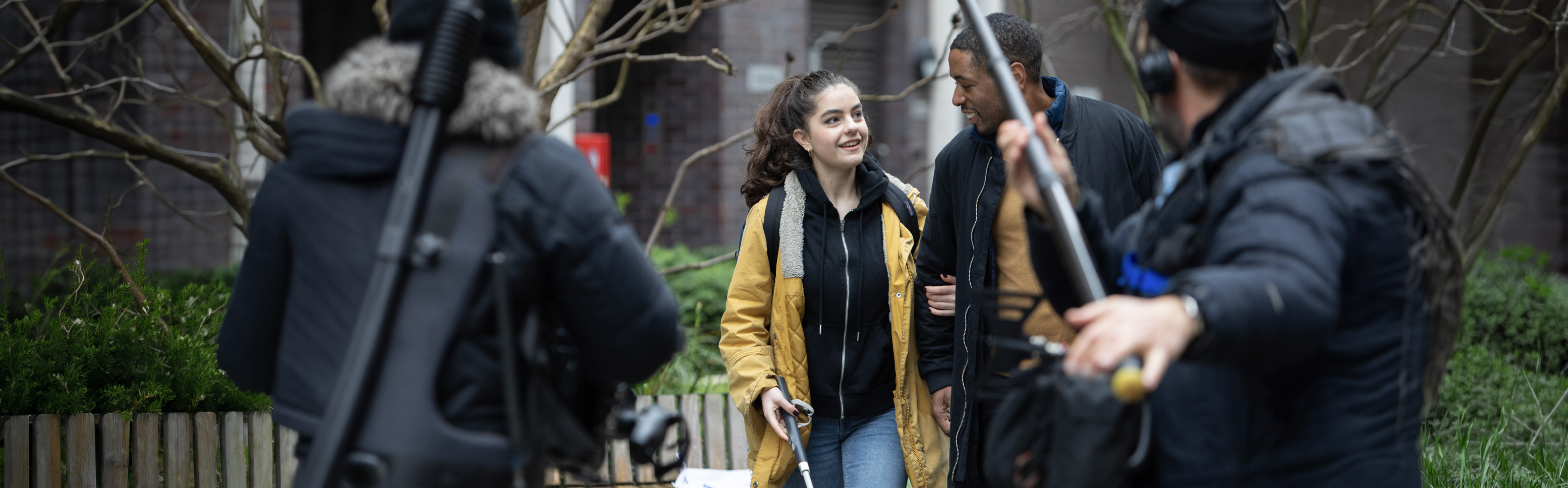 Behind the scenes during filming of Nyla&rsquo;s Journey: a smiling young woman stands outdoors while a film crew with cameras and a boom microphone gathers around her.