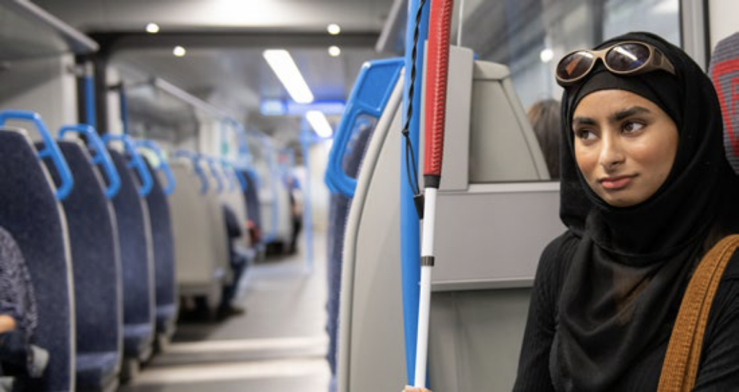 A woman seated on public transport holds a white mobility cane. She wears a black headscarf and has sunglasses resting on her head. Rows of blue-handled seats and passengers are visible in the background.