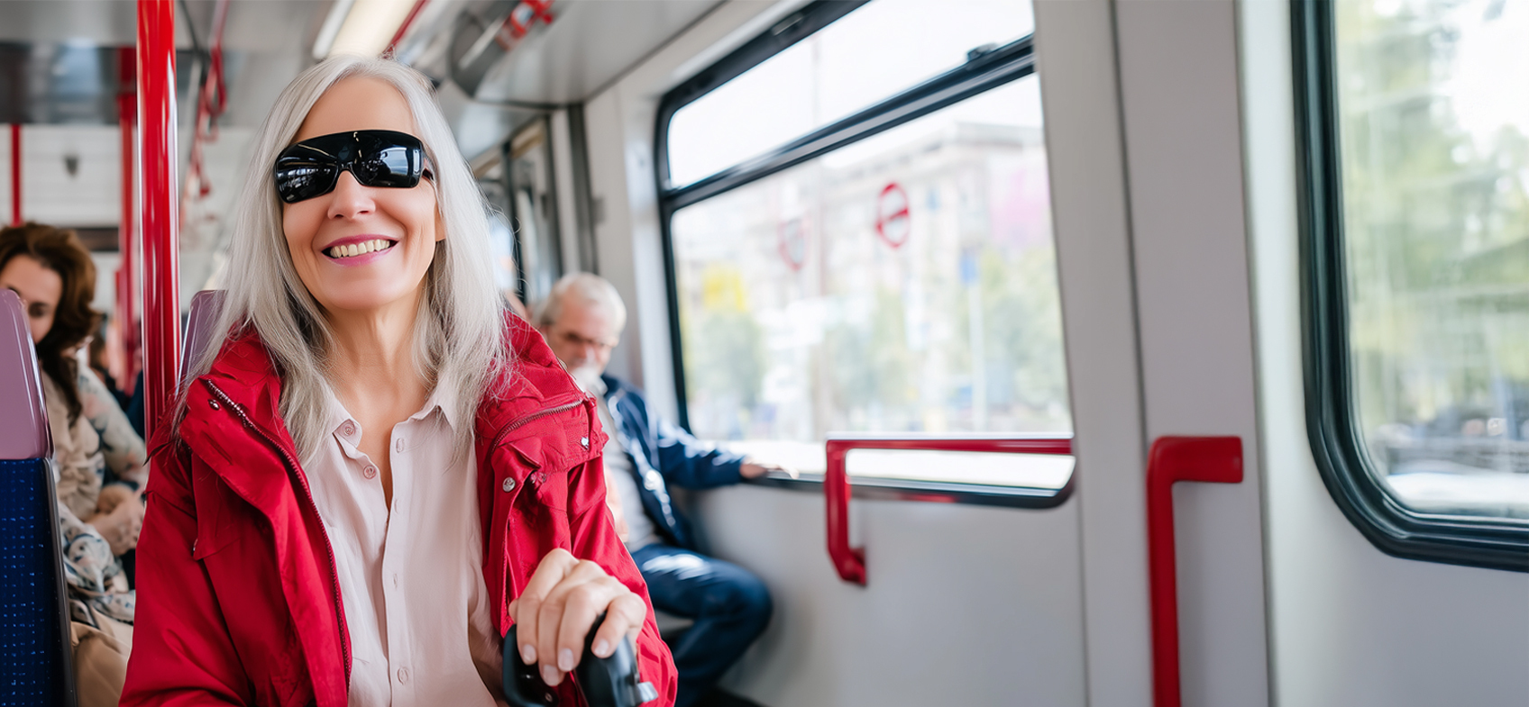 An older woman smiles while seated on public transport. She wears dark wraparound sunglasses and a red jacket, holding a mobility cane in one hand. Other passengers sit in the background and daylight comes through the window beside her.
